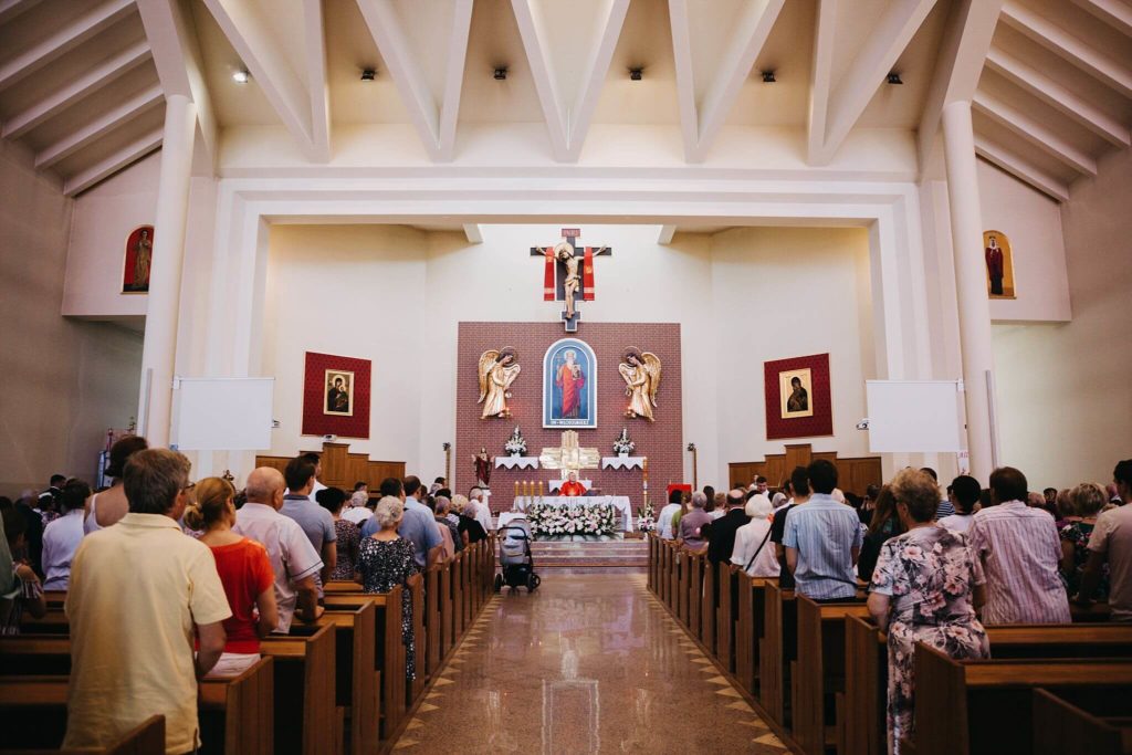 Zdjęcie przedstawia Chrzest Święty w Warszawie w kościele Św. Włodzimierze w Warszawie. Kadr jest szeroki. W centrum ukazuje ołtarz, a po lewej i prawej stronie ludzi stojących w ławkach.